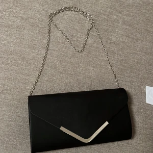 Svart kuvertväska med kedja - Elegant svart kuvertväska med en stilren kedja som axelrem. Väskan har en metallisk detalj på framsidan och ett mönstrat innerfoder. Väskan har även en liten ficka på insidan.