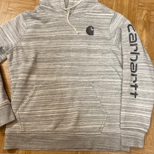 Grå randig hoodie från Carhartt - Säljer en grå randig hoodie från Carhartt med deras logga tryckt på bröstet och ärmen. Hoodien har en känguruficka och justerbar huva med dragsko. Perfekt för en avslappnad stil. Sparsamt använd 