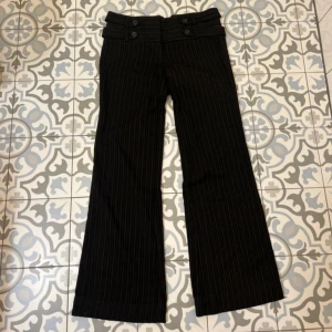 Lågmidjade kostymbyxor - Snygga svarta kostymbyxor med diskreta ränder  och låg midja. Byxorna har en bred linning med dekorativa knappar framtill. Perfekta för en stilren look. Det står stl 10 alltså Medium i byxorna, men skulle absolut säga att de är mer en stl Small. Från topshop. 