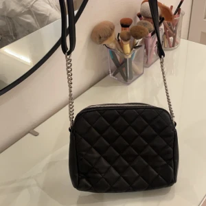 Svart quiltad axelväska - Snygg svart quiltad axelväska med kedjedetaljer på axelremmen. Väskan har en dragkedja upptill och en stilren design som passar perfekt för en trendig look. Perfekt för att bära dina nödvändigheter med stil.