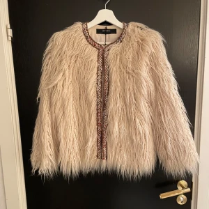 Beige pälsjacka från Zara Basic - Säljer en fluffig beige pälsjacka från Zara Basic. Jackan har en dekorativ kant i olika färger längs öppningen och är perfekt för att ge en lyxig touch till din outfit. Den är långärmad och har en rund halsringning. Den är i storlek S!🪩🥂