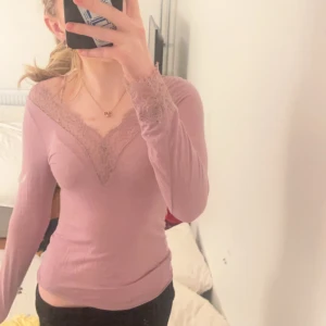 Rosa långärmad topp med spetsdetaljer från Intimissimi - Säljer en söt rosa långärmad topp med v-ringning och spetsdetaljer runt halsen. Perfekt för en stilren och feminin look. Toppen är i ett mjukt och bekvämt material.