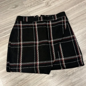 Svart rutig kjol från Trendyol - Snygg svart kjol från Trendyol med ett klassiskt rutigt mönster i vitt och rött. Kjolen har en omlott-design som ger en stilren look. Perfekt för att addera lite mönster till din garderob.