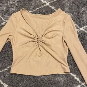 Beige ribbad topp - Säljer en snygg beige ribbad topp med långärmad design och en knutdetalj framtill. Toppen har en v-ringad hals och är perfekt för en stilren look.