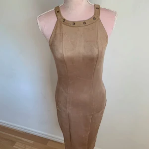 Beige klänning med nitar - Snygg beige klänning med nitar runt halsringningen. Klänningen har en tight passform och är ärmlös, vilket ger en stilren look. Perfekt för en kväll ute eller en speciell tillställning.
