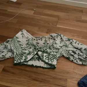 Vit och grön blommig blus - Säljer en söt vit blus med grönt blommönster. Blusen har trekvartsärmar och en volangdetalj framtill. Den är croppad med elastisk nederkant för en bekväm passform.