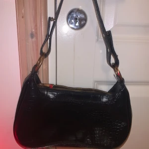 Svart axelväska med krokodilmönster - Snygg svart axelväska i syntet med krokodilmönster och justerbar axelrem. Väskan har en dragkedja upptill och guldfärgade metalldetaljer. Perfekt för att ge din outfit en edgy touch.