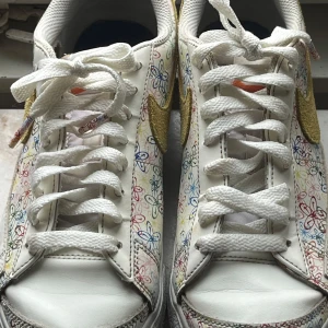 NikeBlazer Low Platform W - Snygga vita Nike sneakers med ett färgglatt blommigt mönster och gyllene swoosh. Skorna har klassisk snörning och en bekväm passform. Perfekta för att ge din outfit en färgglad touch.