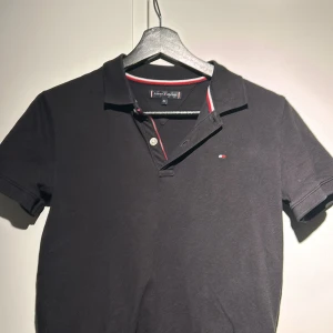 Svart pikétröja från Tommy Hilfiger - Snygg svart pikétröja från Tommy Hilfiger med klassisk krage och knappar. Tröjan har en liten logotyp på bröstet och detaljer i rött och vitt på kragen. Perfekt för en stilren look.