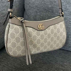 Beige axelväska med monogram - Snygg beige axelväska med ikoniskt monogrammönster och guldfärgad logodetalj. Väskan har en dragkedja och justerbar axelrem. Perfekt för att ge din outfit en lyxig touch.