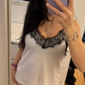 Vit linne med spetsdetaljer - Säljer ett vit linne med svarta spetsdetaljer vid halsringningen. Linnet har tunna axelband och en mjuk, följsam passform. Perfekt för en elegant look. (Aldrig använt)