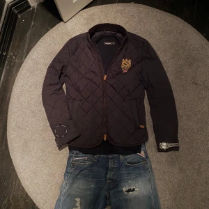 Marinblå quiltad Morris - Snygg marinblå quiltad jacka från Morris med broderad detalj på bröstet. Jackan har en dragkedja framtill och långa ärmar. Perfekt för en stilren look. Jackan är utrustad med dragkedjfickor och innerfickor. Riktigt bra pris och otroligt skick.