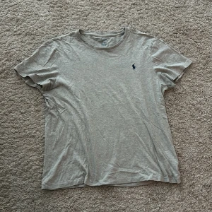 Grå t-shirt från Ralph Lauren - Säljer en beige t-shirt från Ralph Lauren med en broderad logga på bröstet. T-shirten är i bomull och har en normal passform. Perfekt för en avslappnad stil.