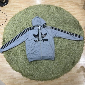 Grå hoodie med dragkedja från Adidas - Säljer en grå hoodie från Adidas med dragkedja och svart logga på framsidan. Tröjan har långa ärmar och klassiska svarta ränder längs ärmarna. Perfekt för en avslappnad stil.