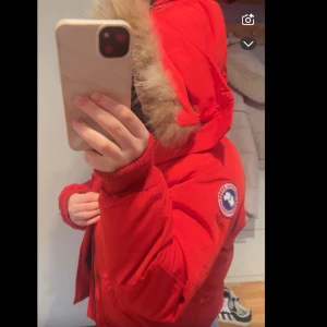 Canada goose jacka - Säljer en snygg röd jacka från Canada Goose med pälsdetaljer på luvan. Jackan har en dragkedja och knappar framtill samt långa ärmar. Perfekt för kalla vinterdagar. Använd en vinter.