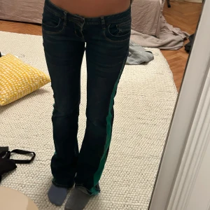 Blå jeans med gröna detaljer - Snygga blå jeans med en unik grön rand längs sidorna. De har en låg midja och en bootcut-stil. Jag är 178 och innebenslängd 89, midjemått 39 men de går att ha ett större midjemått 