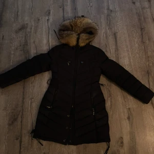 Svart dunjacka med pälsdetalj från Hollies - Snygg svart dunjacka från Hollies med äkta pälsdetalj på huvan. Jackan har dragkedja och knappar framtill samt  fickor med dragkedja. Även leopard material på jackans insida. Nypris för jackan är ca 3000kr