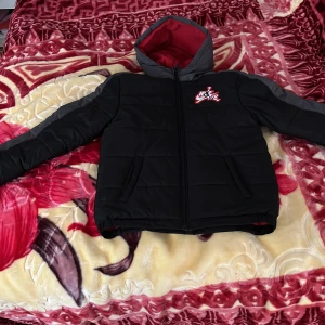 Svart pufferjacka från Nike - Säljer en svart pufferjacka från Nike/Jordan med röd insida och broderad logga på bröstet. Jackan har en dragkedja framtill och långa ärmar. Perfekt för kyliga dagar. Som ni kan se har jag ej använt denna jacka eftersom den var för liten.