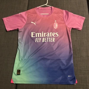 Rosa och blå fotbollströja från Puma - Snygg fotbollströja från AC Milan, med Puma i en cool gradient av rosa och blått. Tröjan har korta ärmar och tryck med 'Emirates Fly Better' på framsidan och 'wefox' på baksidan. Perfekt för fotbollsfans som vill sticka ut på planen!
