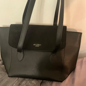 Svart axelväska  - Snygg svart axelväska från Art Street London med stilren design. Väskan har en praktisk dragkedja på baksidan och rymligt inre med flera fack. Perfekt för att bära med sig det nödvändigaste med stil. Aldrig använd