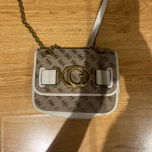 Beige axelväska från Guess - Säljer en snygg beige axelväska från Guess med ett mönster av logotyper och en stor guldlogga på framsidan. Väskan har en justerbar axelrem med kedjedetaljer och stängs med lock. Perfekt för att ge din outfit en lyxig touch!