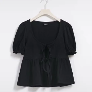 Svart blus med knytning - Säljer en svart blus med korta puffärmar och knytning framtill. Blusen har en luftig passform och är perfekt för både vardag och festliga tillfällen.