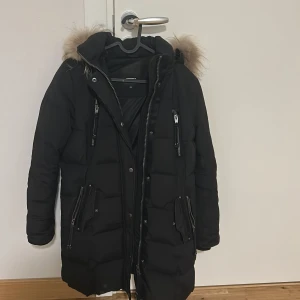 Svart dunjacka med pälsdetalj - Snygg svart dunjacka med pälsdetalj på huvan. Jackan har dragkedja och knappar framtill samt flera praktiska fickor med dragkedjor. Perfekt för kalla dagar med sin långa design och varma material.