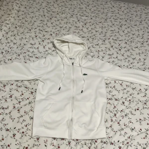 Vit hoodie från Lacoste - Säljer en stilren vit hoodie från Lacoste med dragkedja och huva. Tröjan har långa ärmar och den klassiska Lacoste-loggan på bröstet. Perfekt för en avslappnad look.