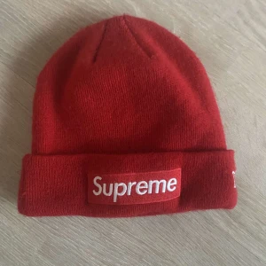 Röd mössa från Supreme - Snygg röd mössa från Supreme med broderad logga framtill och texten 'World Famous' på baksidan. Tillverkad i mjukt material för en bekväm passform. Perfekt för att hålla värmen med stil.