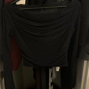 Svart glittrig topp - Helt ny snygg svart glittrig topp med långa ärmar och en elegant drapering framtill. Perfekt för en kväll ute eller en festlig tillställning. Toppen har en åtsittande passform som framhäver figuren.