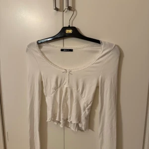 Vit långärmad topp från Gina Tricot - Säljer en söt vit långärmad topp från Gina Tricot. Toppen har spetsdetaljer vid halsringningen och en liten rosett framtill. Den är croppad med volangdetaljer vid nederkanten. Perfekt för en stilren look.