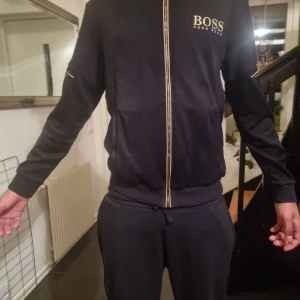 Svart lång armada tröja från Hugo Boss - Snygg svart lång armad från Hugo Boss med gulddetaljer. Jackan har dragkedja och långa armar. Perfekt för en stilren look.