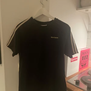 Wales Bonner  - Säljer en svart t-shirt från Adidas med klassiska vita ränder längs ärmarna och en liten logga på bröstet. Perfekt för en sportig look eller avslappnad stil. Passar bra till jeans eller träningsbyxor.
