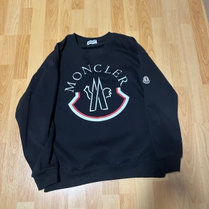 Svart sweatshirt från Moncler - Snygg svart sweatshirt från Moncler med stort tryck på framsidan och broderad logga på ärmen. Perfekt för en stilren och avslappnad look.