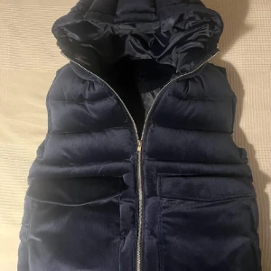 Mörkblå quiltad väst med huva från Vedoneire - Snygg mörkblå quiltad väst från Vedoneire med dragkedja och praktiska fickor. Västen har en mysig huva och är perfekt för kyligare dagar. Den är stilren och lätt att matcha med olika outfits.