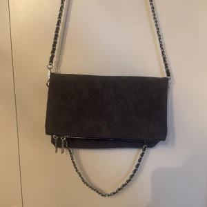 Svart axelväska med kedjedetaljer - Snygg svart axelväska med kedjedetaljer och dragkedja. Finns två fack och även ett fack på baksidan. Två kedjor, så man kan knäppa av det långa om man endast vill ha kort och tvärtom! 