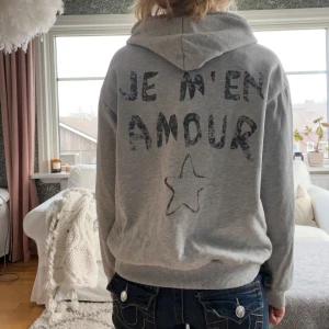 Grå hoodie från Gina Tricot - Säljer en grå hoodie från Gina Tricot Young med texten 'JE M'EN AMOUR' och en stjärna tryckt på både fram- och baksidan. Hoodien har en klassisk design med ribbade muddar vid ärmslut och nederkant. Den är väl använd och väldigt nopprig. Saknas även metallgrej på snöret vid luvan, se bild ovan.