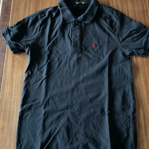 Ralph lauren pique - Snygg svart pikétröja från Ralph Lauren med klassisk krage och korta ärmar. Tröjan har en liten röd logotyp broderad på bröstet. Perfekt för en stilren look.