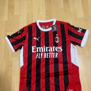 Ac milan fotbollströja - Snygg AC Milan fotbollströja från Puma i röd och svart med vita detaljer. Tröjan är kortärmad och har klubbens emblem på bröstet samt sponsorlogga. Perfekt för fans som vill visa sitt stöd på matchdagar!