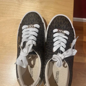 Bruna sneakers från Michael Kors storlek 34 - Snygga bruna sneakers från Michael Kors med vitt snörning och mönstrad yta. Skorna har en klassisk design med en vit sula och Michael Kors-logga på sidan. Perfekta för en stilren look. Aldrig använda. Storlek:34. Köpte för 589 men kan sälja för 530 kronor.✨