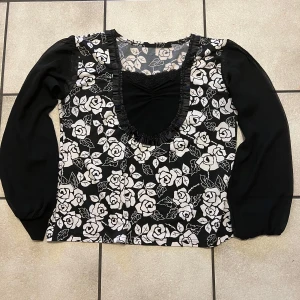 Svart och vit blommig blus storlek M - Snygg svart blus med vita rosor och långa ärmar. Blusen har en v-ringad design med volangdetaljer runt halsen. Perfekt för en stilren look.