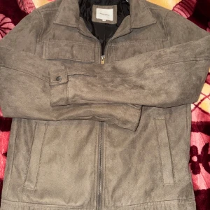 Grå mockajacka från Jack & Jones - Snygg grå mockajacka från Jack & Jones med dragkedja framtill. Jackan har en klassisk krage och två bröstfickor med lock. Perfekt för en stilren look.