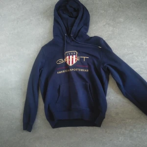 Mörkblå hoodie från Gant - Säljer en snygg mörkblå hoodie från Gant med deras klassiska logga och texten 'American Sportswear' på framsidan. Tröjan har en praktisk magficka och justerbar huva. Perfekt för en avslappnad stil.