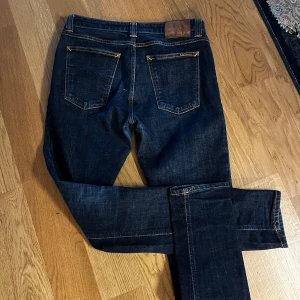 Mörkblå jeans från Nudie Jeans - Snygga mörkblå jeans från Nudie Jeans med klassisk femficksdesign och kontrastsömmar. Jeansen har en smal passform och är perfekta för en stilren look. Märkeslogga broderad på bakfickan. Priset är inte hugget i sten!