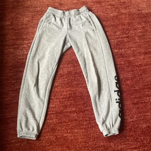 Grå mjukisbyxor från Adidas - Snygga grå mjukisbyxor från Adidas med elastisk midja och muddar vid bensluten. Byxorna har en svart Adidas-logga längs benet och är perfekta för en avslappnad stil. Har en liten fläck på ena benen som syns på bild 3. 