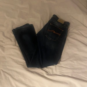 Mörkblå jeans från Nudie Jeans - Snygga mörkblå jeans från Nudie Jeans med klassisk femficksdesign och kontrastsömmar. De har en knappgylf och en stilren passform som passar perfekt till vardags. Märkeslogga på bakfickan ger en extra touch. Moddelen heter slim Jim 