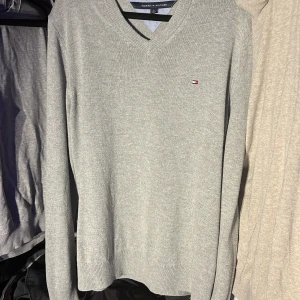 Grå tröja från Tommy Hilfiger - Säljer en stilren grå tröja från Tommy Hilfiger med v-ringning och diskret logga på bröstet. Tröjan är långärmad och tillverkad i en mjuk blandning av bomull och syntetmaterial. Perfekt för en avslappnad och snygg look.