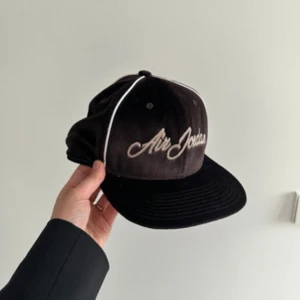 Svart keps från Air Jordan - Snygg svart keps från Air Jordan med vit broderad logga framtill. Kepsen har en klassisk design med vit piping längs panelerna och justerbar passform. Perfekt för en sportig look eller som en stilren accessoar.