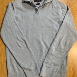 Ljusblå tröja från Tommy Hilfiger - Snygg ljusblå tröja från Tommy Hilfiger i premium bomull. Tröjan har en dragkedja vid halsen och ett diskret broderat logotypmärke på bröstet. Perfekt för en stilren look.