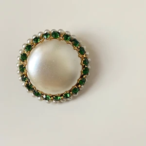 Elegant brosch med pärlor och gröna stenar - Snygg vintage brosch med en stor vit pärla i mitten, omgiven av gnistrande gröna stenar och en rad av små vita pärlor. Baksidan har ett vackert mönster i guld. Perfekt för att ge en touch av elegans till din outfit. 